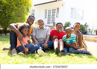 Three Generation African American Family Spending Time In Their Garden On A Sunny Day, Sitting On A Lawn And Smiling.