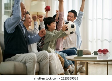 three generation asian family sitting on couch at home celebrating goal and victory while watching live broadcasting of football match - Powered by Shutterstock