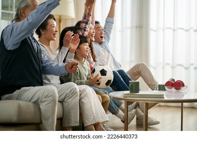 three generation asian family sitting on couch at home celebrating goal and victory while watching live broadcasting of football match - Powered by Shutterstock