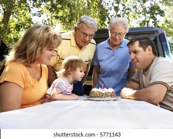 Three Generation Caucasian Family Seated At Picnic Table Celebrating Female Child's Birthday With Cake.