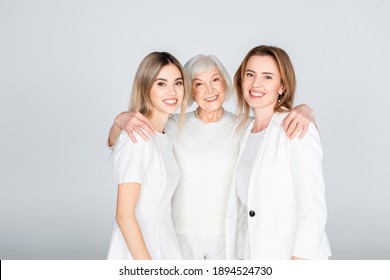 three generation of cheerful women smiling while looking at camera and hugging isolated on grey