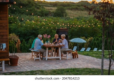 Three Generation Family Having A Garden Party In The Summer. They Are Sitting At The Dining Table In The Backyard And Having Dinner