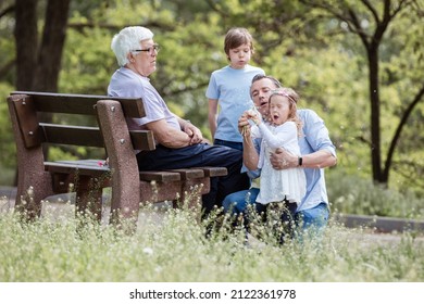 Three Generation Family In Summer Park: Grandfather Sitting On Bench, Father, And Grandchildren. Father And Daughter Blowing Dandelion Seeds.