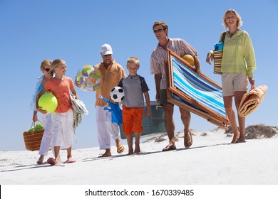 Three Generation Family Walking On Sandy Summer Vacation Beach Carrying Deckchair Bags Towels And Balls