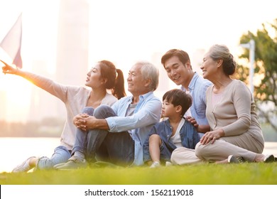 Three Generation Happy Asian Family Sitting On Grass Enjoying Good Time At Dusk Outdoors In Park