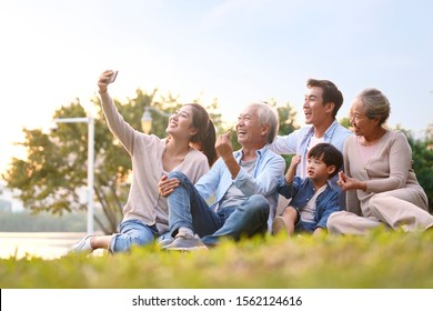 three generation happy asian family sitting on grass taking a selfie using mobile phone outdoors in park - Shutterstock ID 1562124616