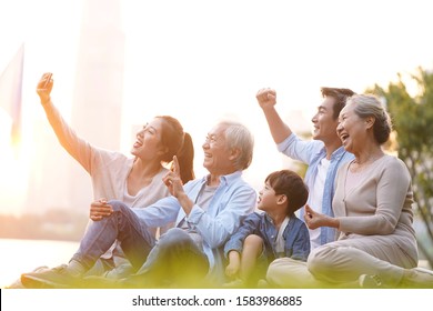 Three Generation Happy Asian Family Sitting On Grass Taking A Selfie Using Mobile Phone Outdoors In Park