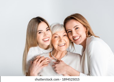 three generation of positive women smiling while looking at camera and hugging isolated on grey
