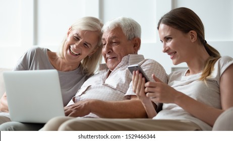 Three generations family with gadgets at home, seated on sofa adult daughter with smartphone and elderly parents using computer, different age people users and modern wireless technology usage concept - Powered by Shutterstock