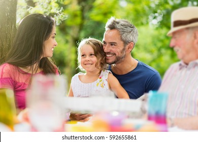 Three Generations Family Gathered To Lunch In The Garden In Summer, They Having Fun Sitting Around A Picnic Table. Shot With Flare