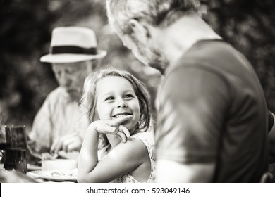 Three Generations Family Gathered To Lunch In The Garden In Summer, Focus On A Beautiful Little Girl Looking At Her Dad. Black And White