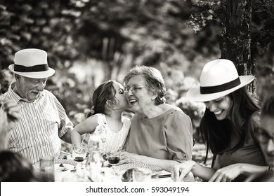 Three Generations Family Having Lunch In The Garden, A Little Girl Whispers A Secret In Her Grandmother's Ear. Black And White