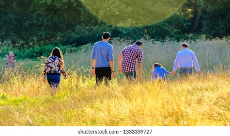 Three Generations Of Family Walking Through Tall Grass In Park At Sunset In Fall; Woods In Background