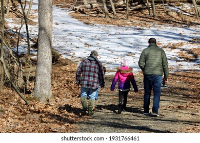 Three Generations Hiking Through The Woods On A Snow Covered Path.  Grandfather, Father, And Daughter Are Walking Alongside Each Other Enjoying The Nature Surrounding Them.