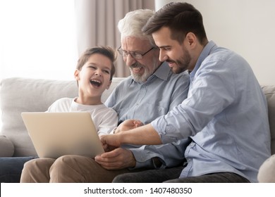 Three generations of men sit on couch having fun watching funny video on laptop together, happy little boy child relax with father and grandfather enjoy movie on computer, laughing resting at home