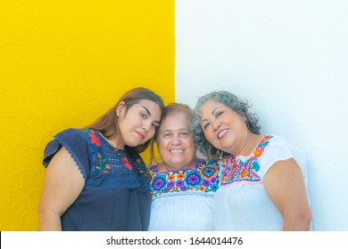 Three generations of Mexican women smiling, granddaughter and daughter leaning on the mother in floral print blouses looking at the camera with a yellow and white background.