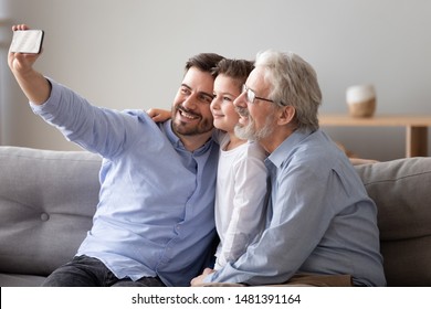 Three Generations Relatives People Men Sitting On Couch In Living Room, Grandad Grown Up Son Grandkid Using Smart Phone Take Selfie Photo Smiling Feels Happy Capture The Moment And Parentage Concept