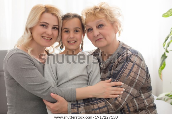 Three\
generations of women. Beautiful woman and teenage girl are kissing\
their granny while sitting on couch at\
home