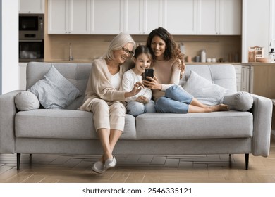 Three generations of women, grandmother, mother, young daughter sitting together on sofa, smiling and looking at smartphone screen, enjoy family moments, internet fun, viewing photos or videos at home - Powered by Shutterstock