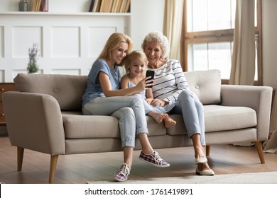 Three generations of women rest on couch have fun watch funny videos on smartphone, happy little girl with old grandmother and mother sit on sofa use modern cellphone making selfie, videocall concept