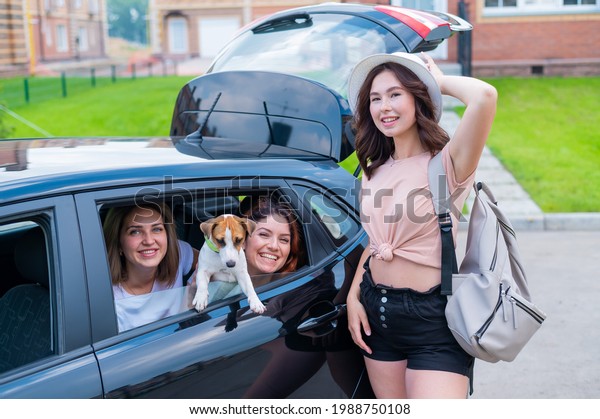 Three girl friends are traveling in a car with a\
dog. Two women are sitting in the back seat with a Jack Russell\
Terrier and looking out of the window, and the third is standing\
nearby and loads