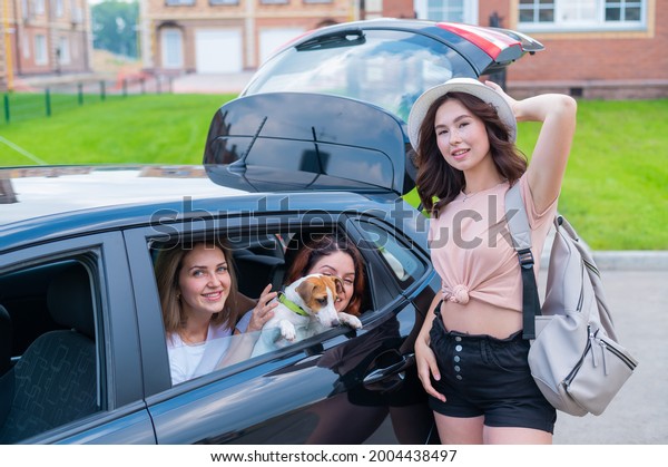 Three girl friends are traveling in a car with a
dog. Two women are sitting in the back seat with a Jack Russell
Terrier and looking out of the window, and the third is standing
nearby and loads
