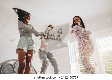 Three Girls Fighting With Pillows Standing On Bed With Feathers Flying Everywhere. Girls Enjoying Pillow Fight Standing On Bed At A Sleepover.