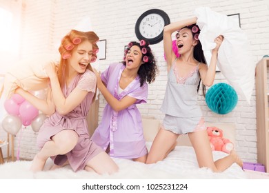 Three Girls In Robes With Curlers In Their Hair Pillow Fight On Bed. They Are Celebrating Women's Day March 8.