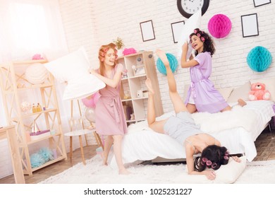 Three Girls In Robes With Curlers In Their Hair Pillow Fight On Bed. They Are Celebrating Women's Day March 8.