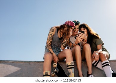 Three Girls Sitting At Skate Park Using Smartphone And Smiling. Female Friends Sitting On Skate Park Ramp With Mobile Phone.
