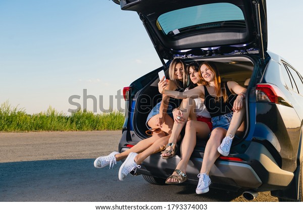 Three girls take a selfie\
together. Blonde, brunette and red haired young women sit down and\
relax at vehicle back, talking during road adventure trip in\
nature.
