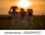 Three girls with wreaths of flowers on their heads stand in the rays of the sunset. Slavic girls in long white dresses look at the sun. Pagan holiday Ivan Kupala. Summer evening