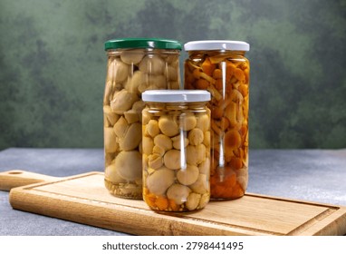 Three glass jars with different types of pickled mushrooms. Including small button mushrooms and wild honey agaric. Standing on a rustic wooden cutting board