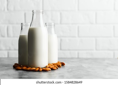 Three glass reusable bottles with almond milk and almond nuts on a marble countertop, a kitchen with a white brick wall.