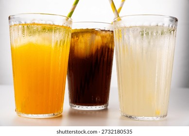 Three glasses filled with cold, refreshing drinks like orange juice, sparkling cola, and lemonade, served with ice and striped straws