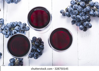 Three Glasses Of Red Wine And Blue Grapes On White Wooden Table Background