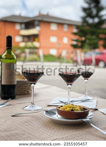 Three glasses of red wine, bottle of wine and small plate of paella served on table outdoor terrace. Vertical urban blurred background