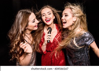 Three Gorgeous Young Women In Festive Dresses Singing Karaoke  Isolated On Black