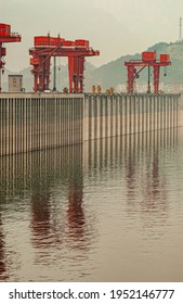 Three Gorges Dam, China - May 6, 2010: Yangtze River. Red Flood Control Door Cranes On Top Of Brown Concrete Dam Reflected In Greenish Water Under Foggy Morning Sky.