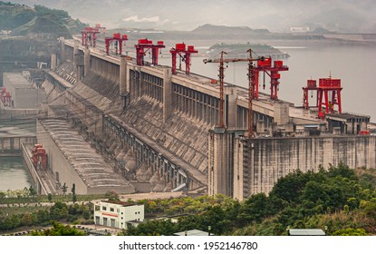 Three Gorges Dam, China - May 6, 2010: Yangtze River. Down River Side Of Dam With Hydroelectric Plant. Red Flood Control Door Lifts On Top Under Foggy Morning Sky. Plenty Of Electric Cables.