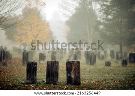 Three gravestones standing in a foggy graveyard during the fall. Focus is on front headstone.