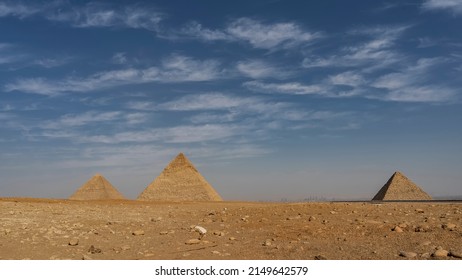 The three great pyramids of Giza - Cheops, Khafre, Menkaur - against the blue sky and clouds. In the foreground is a rocky-sandy desert. Silhouettes of Cairo buildings on the horizon. Egypt
