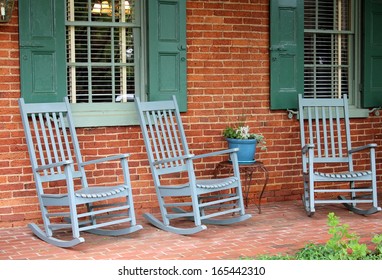 Three Green Adirondack Rocking Chairs Sit Under Rural Country Brick-front Home With Shuttered Windows On Old, Weathered Front Porch Invite Friends And Family To Come Sit Awhile.