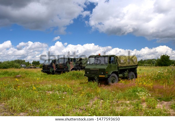 Three green camo military transport trucks
standing in the middle of a field, pastureland or meadow with some
tanks at the back supporting them seen on a cloudy summer day on a
Polish coutryside