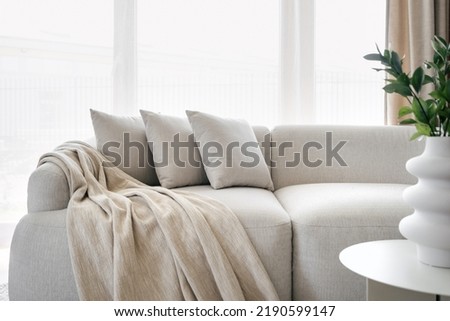 Three grey pillows and blanket on grey soft sofa in living room with panoramic windows. Hypoallergenic concept. Relax idea. Home comfort. House interior details. Hotel room interior design
