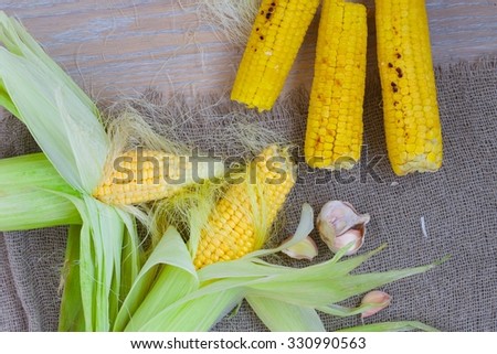 Three grilled and two fresh picked up corn cobs on a sackcloth.Cloves of garlic.Background