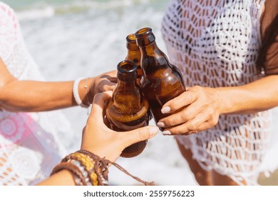 Three hands hold and clink beer bottles together during a toast on a sandy beach with the ocean waves gently lapping in the background and the sky clear and bright blue. - Powered by Shutterstock