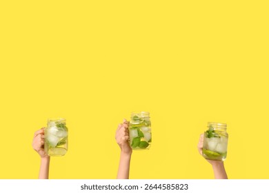 Three hands holding refreshing lime and mint infused water in mason jars, healthy summer beverage on bright yellow background