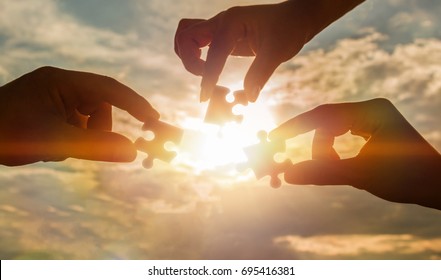 Three hands trying to connect a piece of the puzzle with the sunset background. A puzzle in hand against sunlight. One part of the whole. Symbol of association and communication. Business strategy.