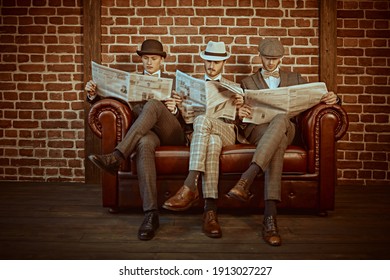 Three Handsome Men In Elegant Suits Sit On A Leather Sofa And Read Newspapers. Newspaper Editorial Office. Retro Style. Men's Fashion.